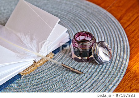 Small journal book and white feather quill lay on a placemat next to an open ink pot Small journal book and white feather quill lay on a placemat next to an open ink pot 97197833