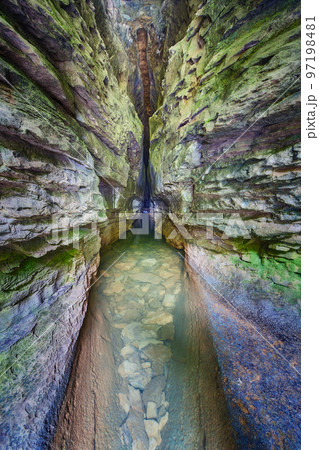 White, green and blue stone walls with a river of clear water leading into a dark and ominous cave 97198481