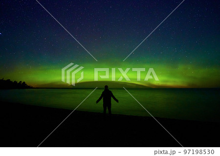 Man on edge of land before water lake ocean or sea with dome of green aurora light mist and tree silhouettes off to side 97198530