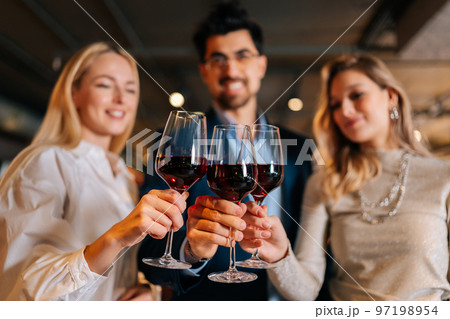Focused on foreground of cheerful man in suit and eyeglasses and two pretty blonde women holding glasses of red wine, standing posing in restaurant. Happy young male and female enjoying nice dinner. 97198954