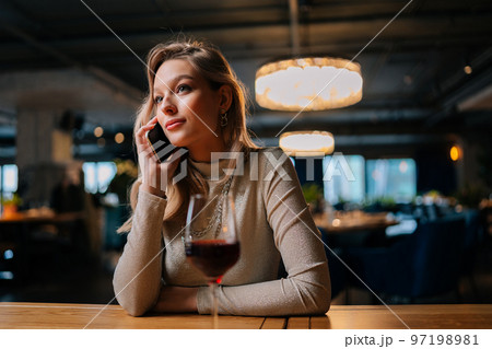 Portrait of bored pretty young woman talking on smartphone with boyfriend who late, sitting in restaurant with glass of red wine. Cute Caucasian female having phone conversation indoors in evening. Portrait of bored pretty young woman talking on smartphone with boyfriend who late, sitting in restaurant with glass of red wine. Cute Caucasian female having phone conversation indoors in evening. 97198981