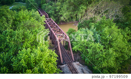 Aerial view above large metal bridge for train tracks over river surrounded by green trees Aerial view above large metal bridge for train tracks over river surrounded by green trees 97199509