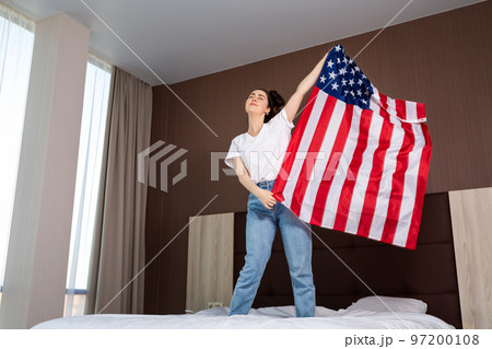 A young happy woman waves an American flag over her bed. Indoor. Bottom view. Concept of the American national holidays and patriotism 97200108
