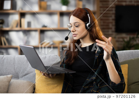 Front view of happy red hair girl in headset studying with laptop at home 97201126