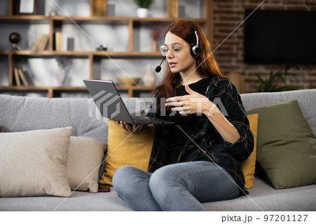 Front view of happy red hair girl in headset studying with laptop at home Front view of happy red hair girl in headset studying with laptop at home 97201127