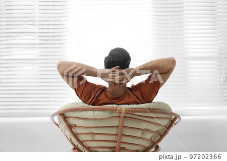 Man relaxing in papasan chair near window at home, back view 97202366