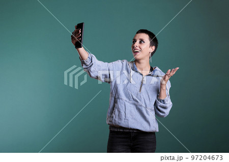 Cheerful caucasian female smiling while talking with remote friend during online videocall meeting on phone, standing in studio with isolated background. Woman chatting with team on videoconference 97204673