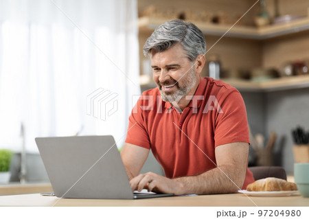 Positive mature man typing on laptop keyboard, kitchen interior Positive mature man typing on laptop keyboard, kitchen interior 97204980