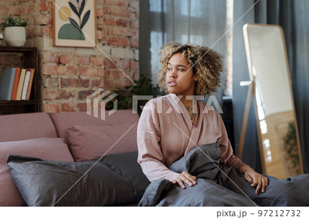 Young relaxed woman in pink pajamas sitting on bed under grey blanket Young relaxed woman in pink pajamas sitting on bed under grey blanket 97212732