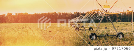Irrigation Machine At Agricultural Field With Young Sprouts, Green Plants On Black Soil. Panoramic View Of Sunset Sky Above Irrigation Pivot. Farming Sprinklers In Field Irrigation, Watering Of Crops 97217066