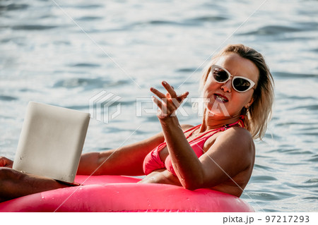 Woman works on laptop in sea. Freelancer, young blond woman in sunglases floating on an inflatable big pink donut with a laptop in the sea at sunset. Freelance, travel and holidays concept. 97217293