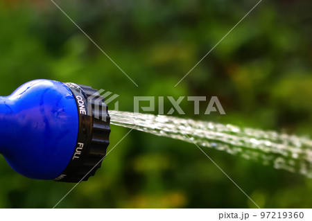 Defocus closeup water garden. Female hand holding blue automatic sprinkler sprays water over brown blurred background. Gardener watering the lawn in the garden. Out of focus 97219360