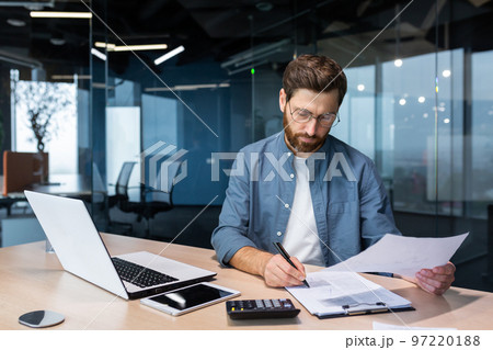 Serious and focused financier accountant on paper work inside office, mature man using calculator and laptop for calculating reports and summarizing accounts, businessman at work in casual clothes. Serious and focused financier accountant on paper work inside office, mature man using calculator and laptop for calculating reports and summarizing accounts, businessman at work in casual clothes. 97220188