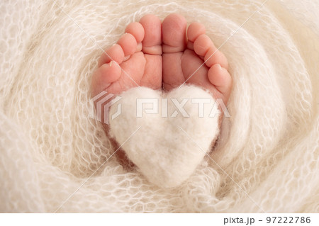 The tiny foot of a newborn baby. Soft feet of a new born in a white wool blanket. Close up of toes, heels and feet of a newborn. Knitted white heart in the legs of a baby. Macro photography. The tiny foot of a newborn baby. Soft feet of a new born in a white wool blanket. Close up of toes, heels and feet of a newborn. Knitted white heart in the legs of a baby. Macro photography. 97222786