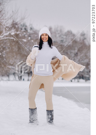 A beautiful girl in a beige cardigan and a white hat with a glass of tea enjoys a snowy embankment by the lake A beautiful girl in a beige cardigan and a white hat with a glass of tea enjoys a snowy embankment by the lake 97223703