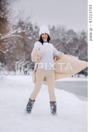 A beautiful girl in a beige cardigan and a white hat with a glass of tea enjoys a snowy embankment by the lake A beautiful girl in a beige cardigan and a white hat with a glass of tea enjoys a snowy embankment by the lake 97223705