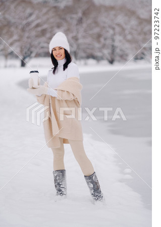 A beautiful girl in a beige cardigan and a white hat with a glass of tea enjoys a snowy embankment by the lake 97223742