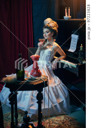 Cocktail evening. Portrait of young beautiful girl in image of medieval person in elegant white dress sitting at the piano. Comparison of eras, beauty, history, art 97223828