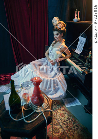 Flirty look. Portrait of young beautiful girl in image of medieval person in elegant white dress sitting at the piano. Comparison of eras, beauty, history, art Flirty look. Portrait of young beautiful girl in image of medieval person in elegant white dress sitting at the piano. Comparison of eras, beauty, history, art 97223916