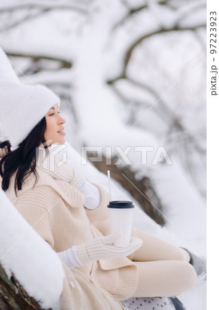A beautiful girl with a beige cardigan and a white hat enjoying drinking tea in a snowy winter forest near a lake 97223923