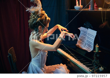 Emotional performance. Portrait of young beautiful woman in image of medieval person sitting at the piano in elegant white dress and playing. Comparison of eras, beauty, history, art 97223957