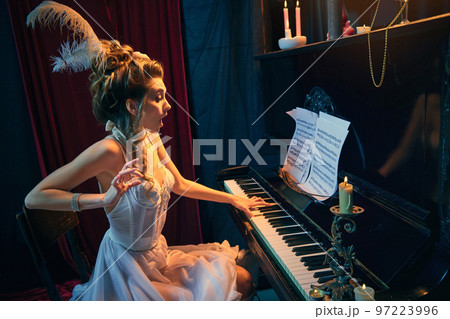 Coquettish look. Portrait of young beautiful woman in image of medieval person sitting at the piano in elegant white dress and playing. Comparison of eras, beauty, history, art 97223996