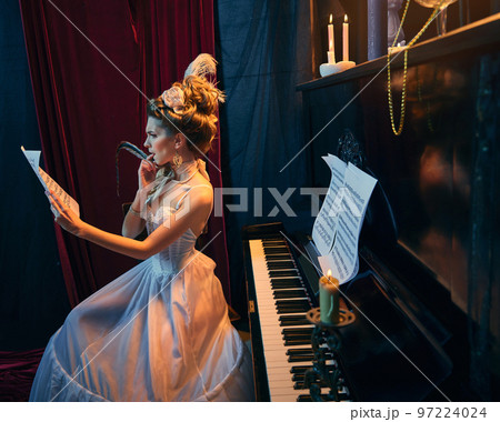 Portrait of young emotive girl in image of royal person, princess sitting at the piano and learning music notes. Music lessons. Comparison of eras, beauty, history, art 97224024