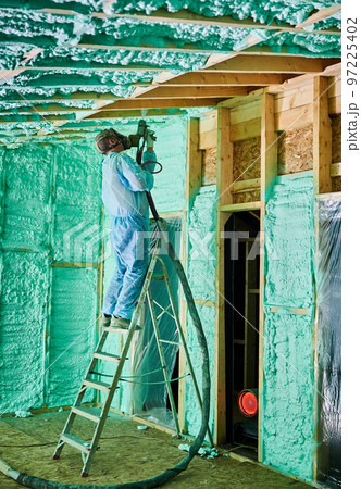 Male builder insulating wooden frame house. Man worker spraying polyurethane foam inside of future cottage, standing on ladder, using plural component gun. Construction and insulation concept. Male builder insulating wooden frame house. Man worker spraying polyurethane foam inside of future cottage, standing on ladder, using plural component gun. Construction and insulation concept. 97225402