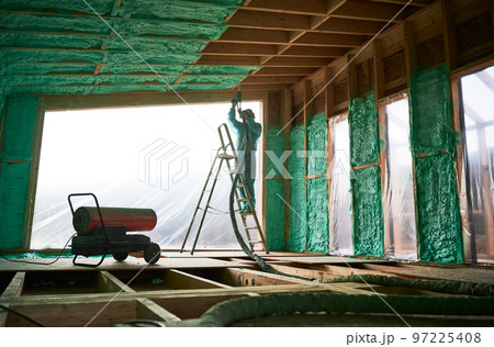 Male builder insulating wooden frame house. Man worker spraying polyurethane foam inside of future cottage, standing on ladder, using plural component gun. Construction and insulation concept. Male builder insulating wooden frame house. Man worker spraying polyurethane foam inside of future cottage, standing on ladder, using plural component gun. Construction and insulation concept. 97225408