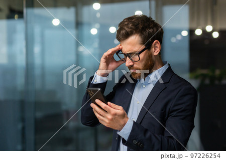 A worried young man businessman holds the phone in his hands, looks. Reading bad news, received a message. Shocked, holds his head. A worried young man businessman holds the phone in his hands, looks. Reading bad news, received a message. Shocked, holds his head. 97226254