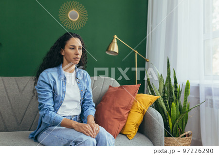 Portrait of sad woman alone at home, pensive Hispanic woman sitting on sofa in living room. Portrait of sad woman alone at home, pensive Hispanic woman sitting on sofa in living room. 97226290