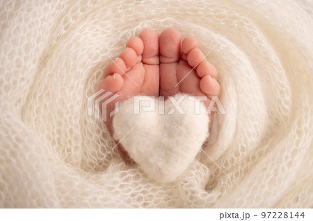 The tiny foot of a newborn baby. Soft feet of a new born in a white wool blanket. Close up of toes, heels and feet of a newborn. Knitted white heart in the legs of a baby. Macro photography.  97228144