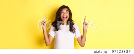 Portrait of amazed, happy african-american girl, smiling and pointing fingers up, standing over yellow background Portrait of amazed, happy african-american girl, smiling and pointing fingers up, standing over yellow background 97228387