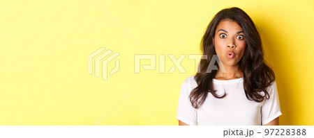 Close-up of amazed african-american girl, gasping and looking at something awesome, standing near copy space over yellow background Close-up of amazed african-american girl, gasping and looking at something awesome, standing near copy space over yellow background 97228388