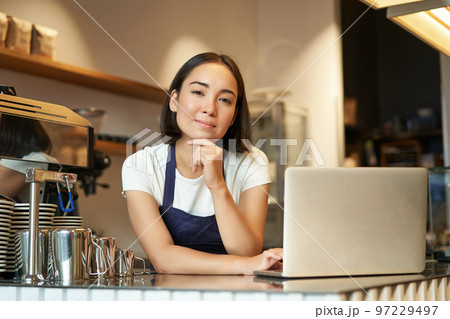 Portrait of smiling korean woman, barista in coffee shop, standing at counter with laptop, smiling and looking confident, self-employed female entrepreneur in her own coffee shop Portrait of smiling korean woman, barista in coffee shop, standing at counter with laptop, smiling and looking confident, self-employed female entrepreneur in her own coffee shop 97229497