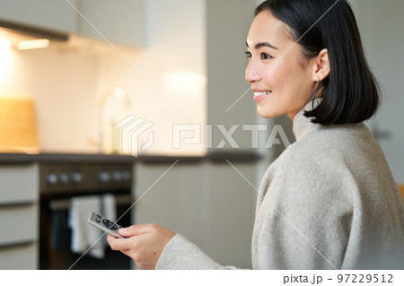 Close up portrait of smiling asian woman watching television, sitting on sofa with remote and switching chanel, looking relaxed Close up portrait of smiling asian woman watching television, sitting on sofa with remote and switching chanel, looking relaxed 97229512