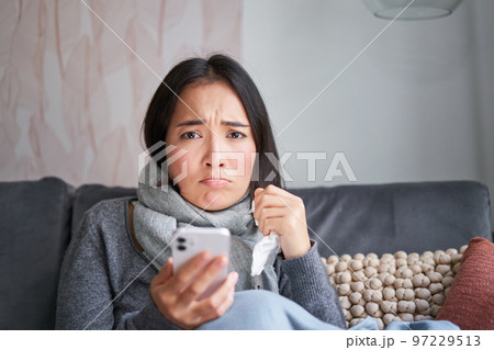 Asian girl with influenza, sits at home, cries and looks upset, holds smartphone, feels unwell and gloomy, wearing warm clothes 97229513
