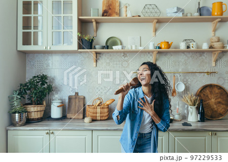 Young beautiful woman at home singing and dancing in the kitchen, cheerful Latin American woman having fun alone with curly hair. Young beautiful woman at home singing and dancing in the kitchen, cheerful Latin American woman having fun alone with curly hair. 97229533