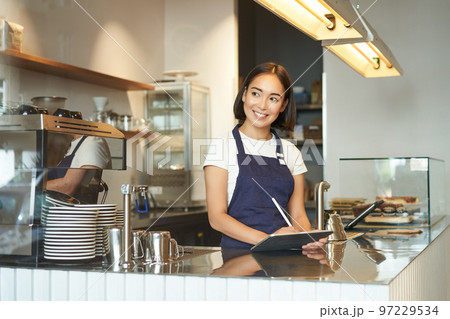 Portrait of beautiful asian girl smiling, barista in cafe working behind counter, using tablet as POS terminal, processing order Portrait of beautiful asian girl smiling, barista in cafe working behind counter, using tablet as POS terminal, processing order 97229534