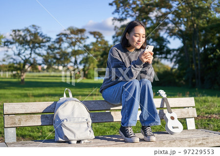 Young woman sitting in park on bench with ukulele, looking at smartphone, reading message on mobile phone and smiling 97229553