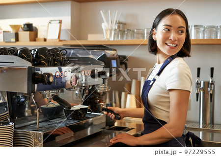 Smiling asian barista girl makes cappuccino with coffee machine, stands behind counter in cafe Smiling asian barista girl makes cappuccino with coffee machine, stands behind counter in cafe 97229555