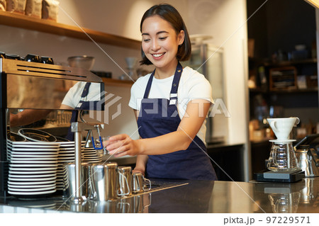 Smiling girl barista in cafe, preparing cappuccino in coffee machine, steaming milk, wearing uniform apron 97229571