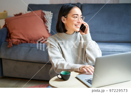 Portrait of smiling asian woman making phone call, working on laptop and having conversation on smartphone 97229573