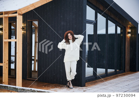 Curly young woman resting on terrace of modern barn house in the mountains. Happy female tourist enjoying winter holidays in new cottage. 97230094