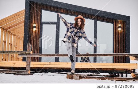 Curly young woman resting on terrace of modern barn house in the mountains. Happy female tourist enjoying winter holidays in new cottage. Curly young woman resting on terrace of modern barn house in the mountains. Happy female tourist enjoying winter holidays in new cottage. 97230095
