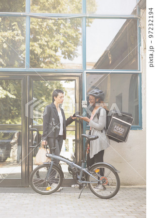 Customer paying for delivery, smartphone mobile wallet contactless payment, nfc technology. African american courier holding pos terminal, standing near office building outdoors 97232194