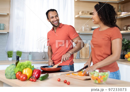 Smiling young black husband and wife in red t-shirts cuts salad, prepares lunch, cooking together in kitchen 97233073