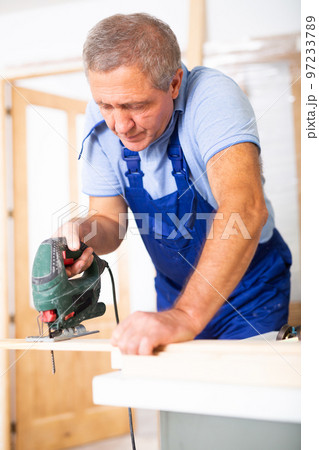 Experienced male worker cuts board with an electric jigsaw in room being renovated 97233789