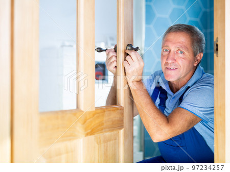 Professional middle-aged repairman in uniform installing a door handle on a door during renovation, indoors Professional middle-aged repairman in uniform installing a door handle on a door during renovation, indoors 97234257
