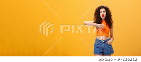 Party for losers I am out. Dissatisfied attractive popular girl with curly hairstyle in red lipstick and cropped top rolling eyes up from irritation and dislike showing thumb down over orange wall 97236212